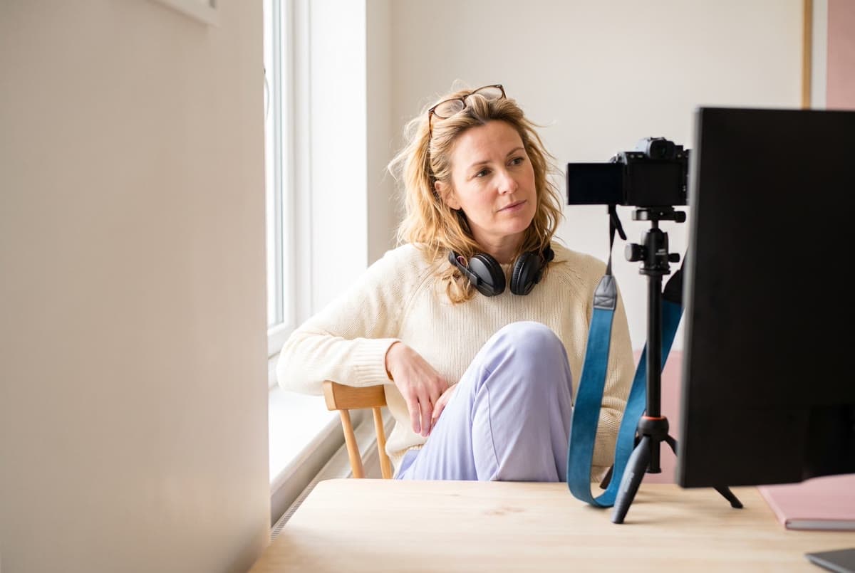Střihačka dokumentárních filmů pracuje u vysokého okna a vedle jejího stolu stojí kamera na stativu.