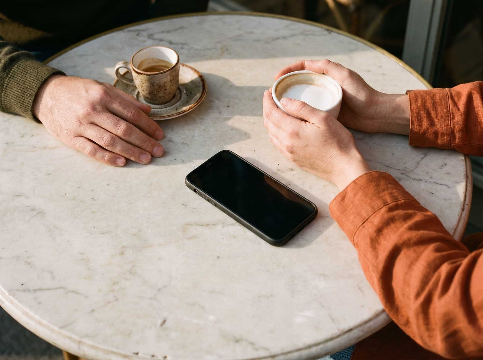 Two people talk over coffee at a small marble café table; a phone lies between their cups, recording.