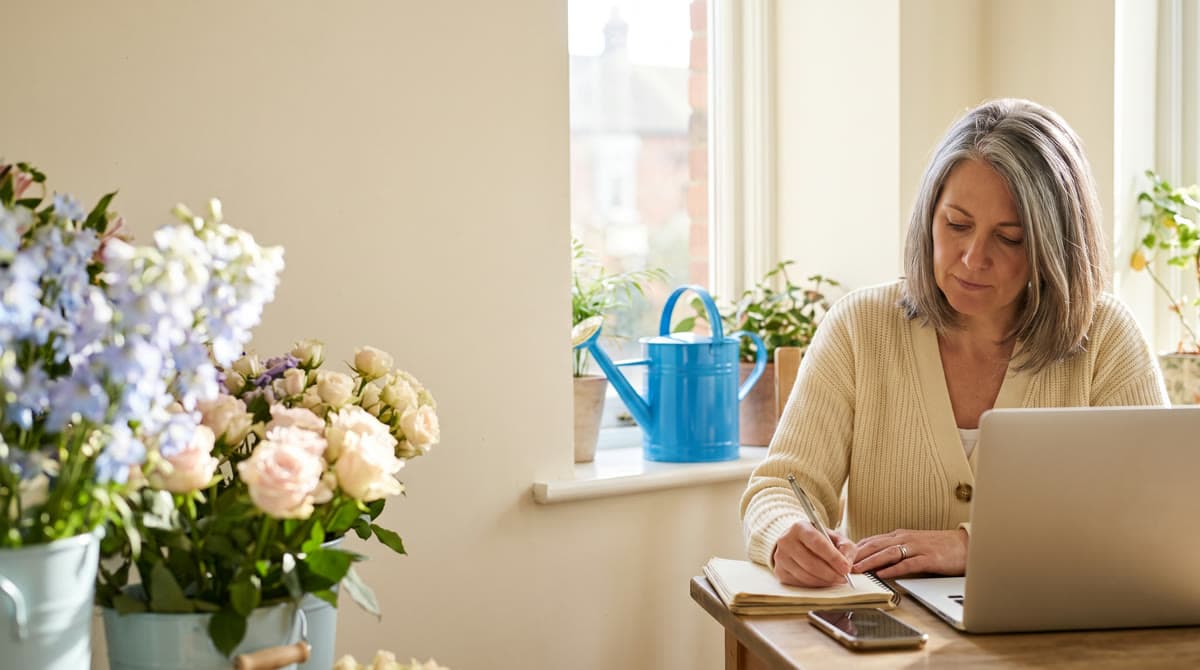 A flower shop owner drafts a post in her notebook in her shop, her laptop beside her.