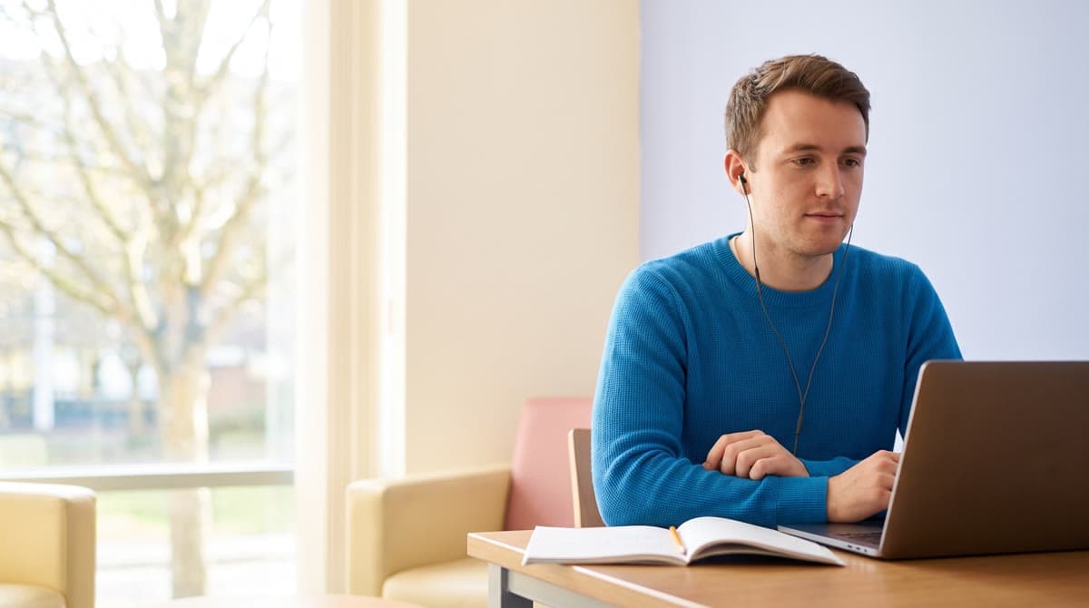 Mladý muž se sluchátky v uších pracuje u notebooku, vedle na stole leží otevřený blok.
