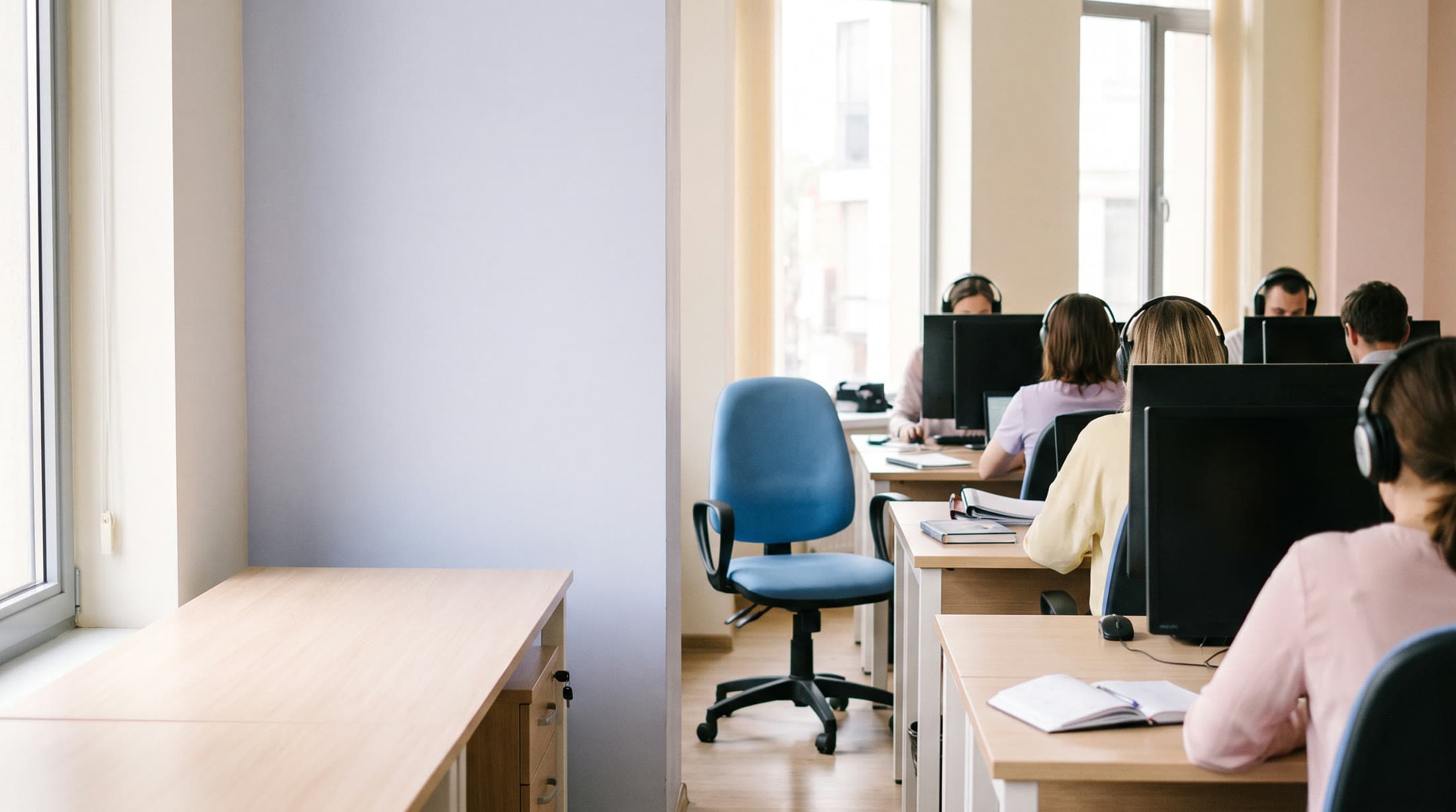 An open-plan office seen from the back of the room: people at light wooden desks with headphones, their monitors facing away, and an empty desk by the window.