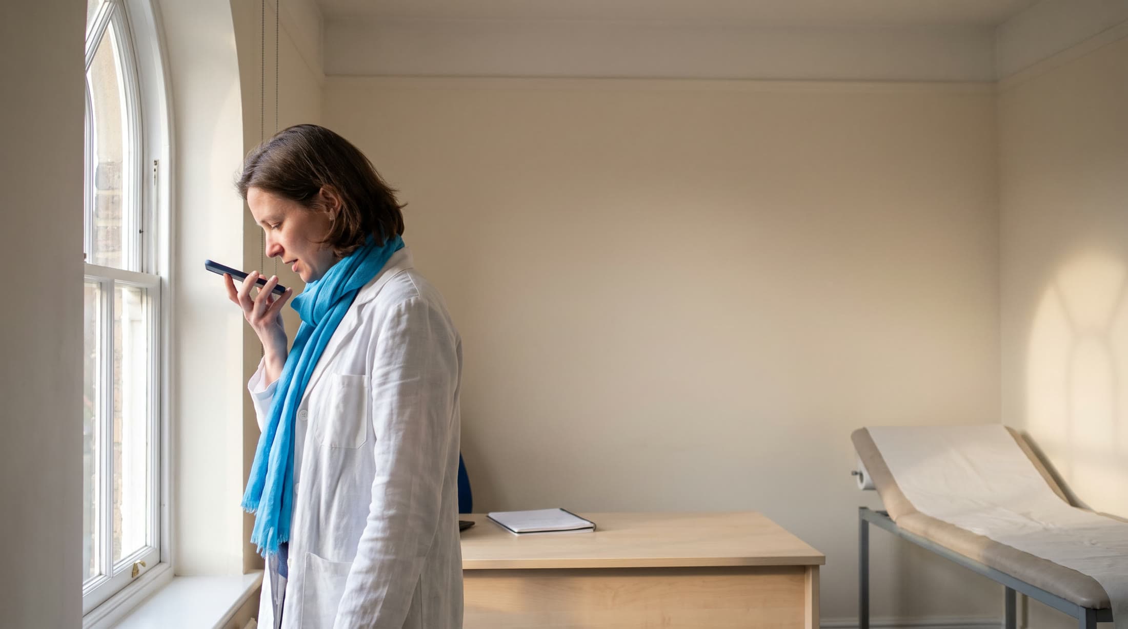 Una professionista sanitaria in camice bianco e foulard blu parla nel telefono davanti alla grande finestra di un ambulatorio vuoto, con una scrivania e un lettino da visita alle spalle.