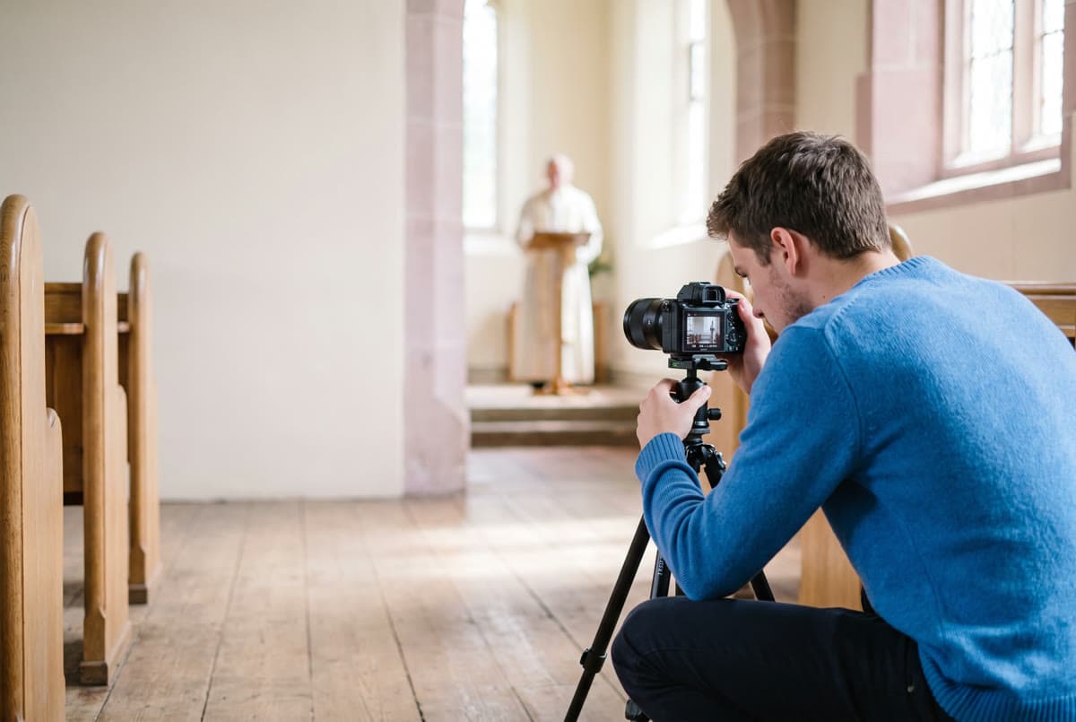 En ung mand sidder på hug i midtergangen i en kirke og stiller et kamera op på et stativ, med prædikanten ved en pult langt bag sig.