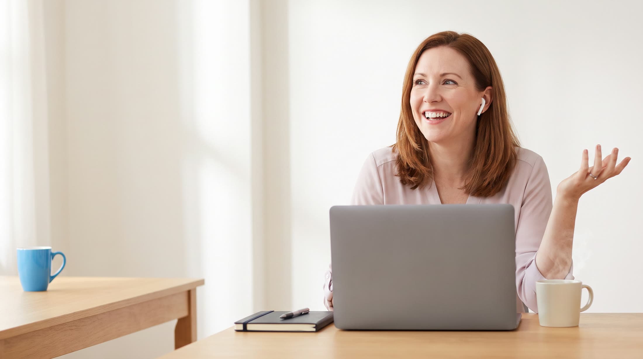 A woman at her desk laughs during a video call on her laptop, an earbud in, a notebook beside her.