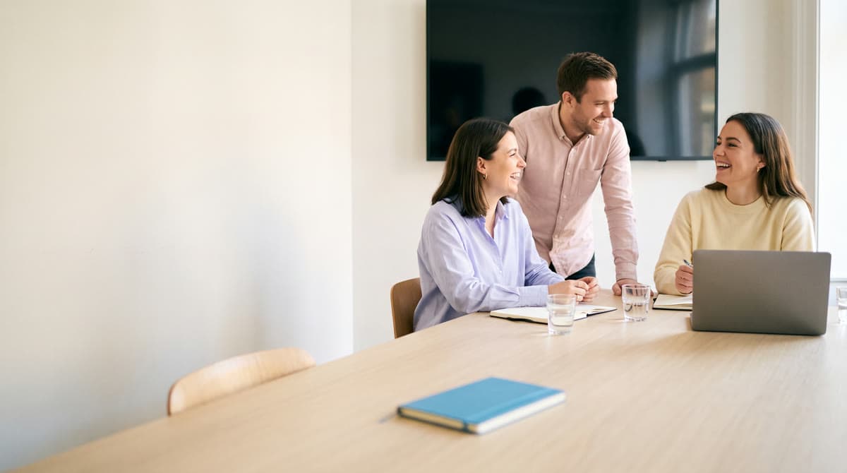 Three colleagues laugh around a meeting room table after a video call, a laptop at the end of the table.
